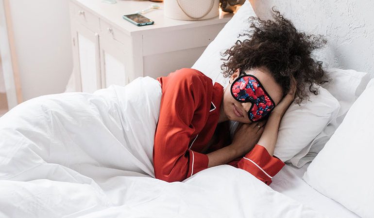 a woman wearing eyemask and red pyjamas sleeping on a bed with white sheets and pillows